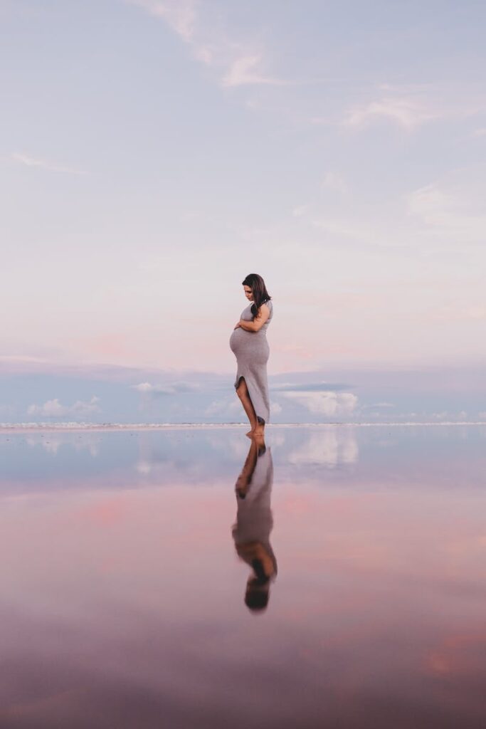 Pregnant woman gracefully stands by the beach during a calming sunset, captured with reflection.