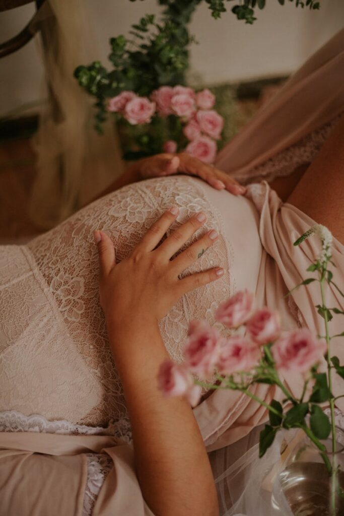 Close-up of expectant mother in lace dress with hand on belly surrounded by pink flowers.